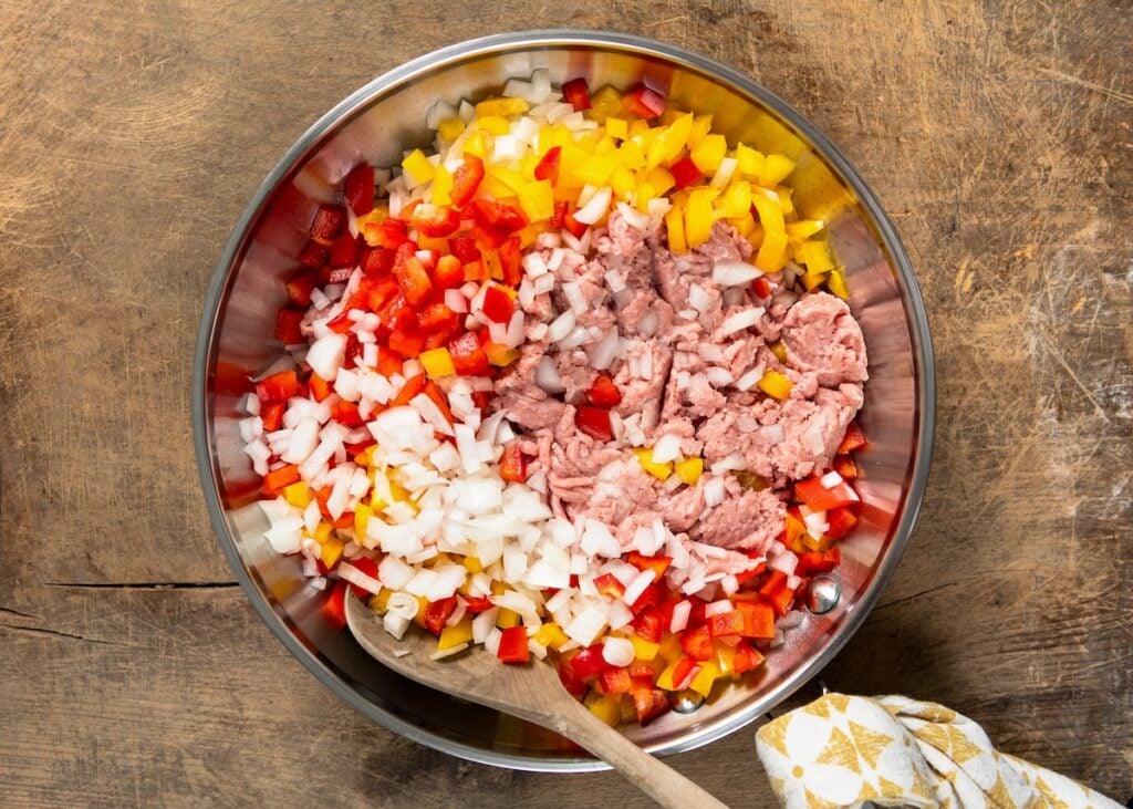 A skillet filled with chopped yellow and red bell peppers, diced onions, and pink ground turkey being stirred with a wooden spoon.