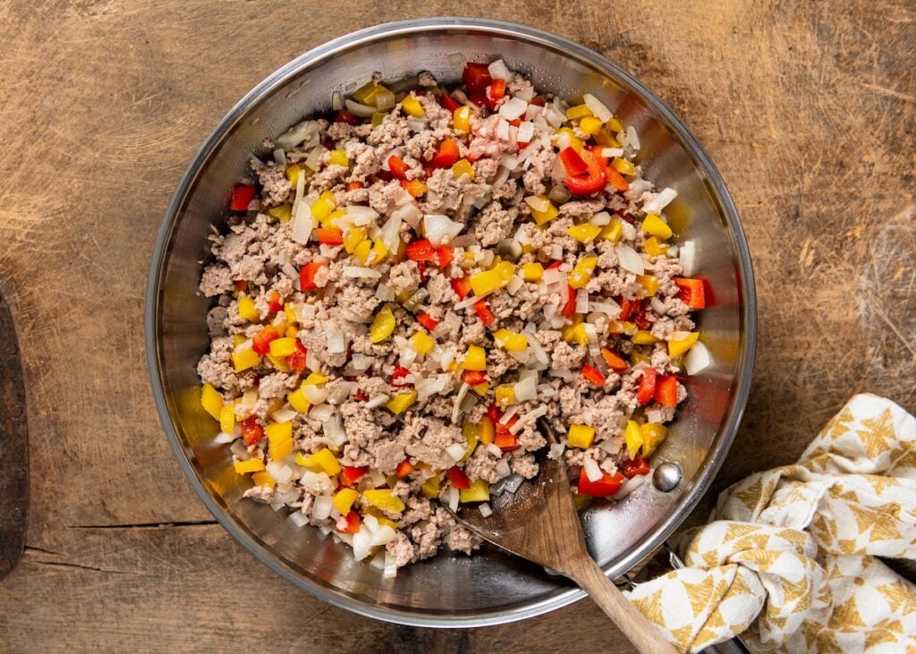 A skillet filled with cooked ground turkey, chopped red and yellow bell peppers, and diced onions, stirred with a wooden spoon.