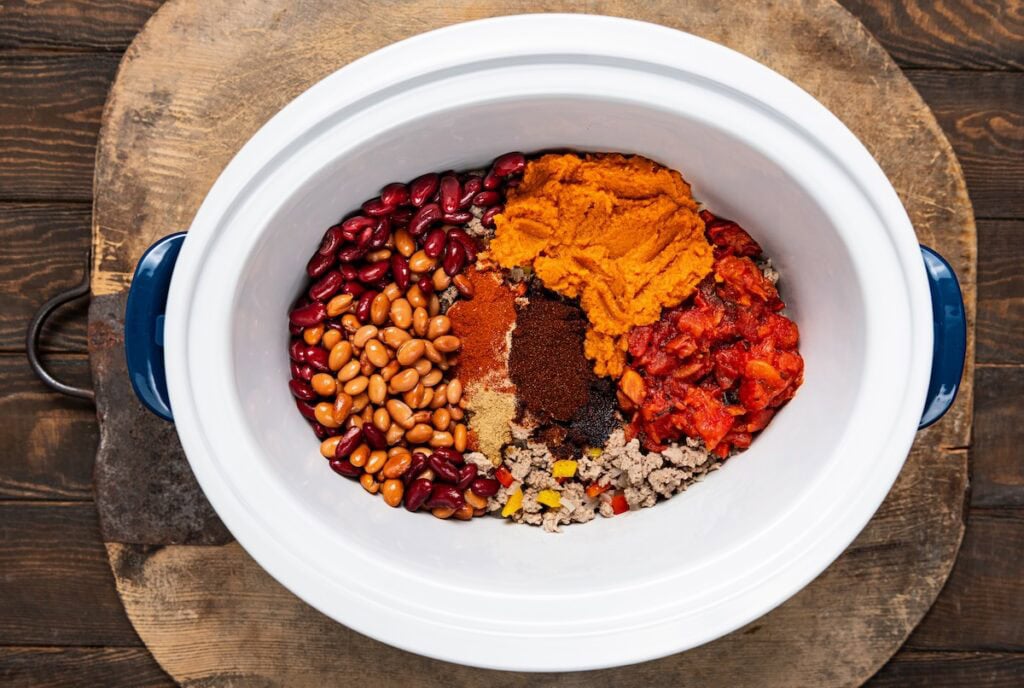 Overhead view of a white slow cooker filled with kidney beans, pinto beans, cooked ground turkey, chopped tomatoes, pumpkin puree, and various spices, all sitting on a wooden surface.
