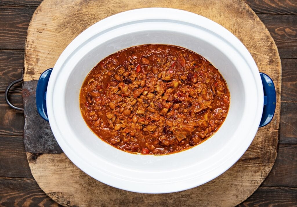 A white crockpot filled with hearty turkey pumpkin chili sits on a round wooden board atop a dark wooden table.