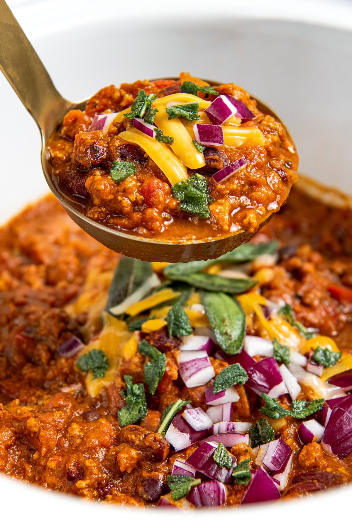 A ladle full of crockpot pumpkin turkey chili topped with shredded cheese, chopped red onions, and herbs is held above a crockpot filled with the same chili and garnishes.