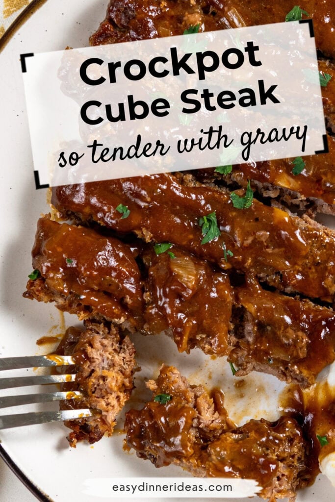 Sliced cube steak covered in brown gravy on a white plate, garnished with chopped parsley. A fork holds a piece of steak. Text over the image reads Crockpot Cube Steak so tender with gravy..