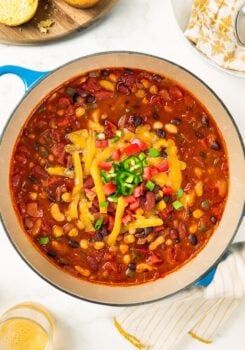 A blue pot of bean chili topped with shredded cheese, diced tomatoes, and sliced jalapeños, with cornbread nearby.