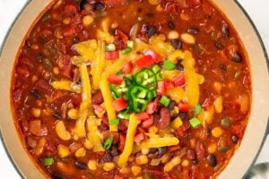A blue pot of bean chili topped with shredded cheese, diced tomatoes, and sliced jalapeños, with cornbread nearby.