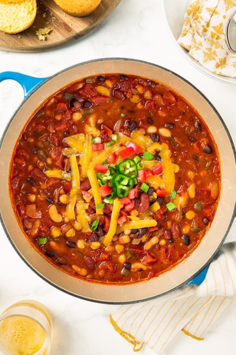 A blue pot of bean chili topped with shredded cheese, diced tomatoes, and sliced jalapeños, with cornbread nearby.