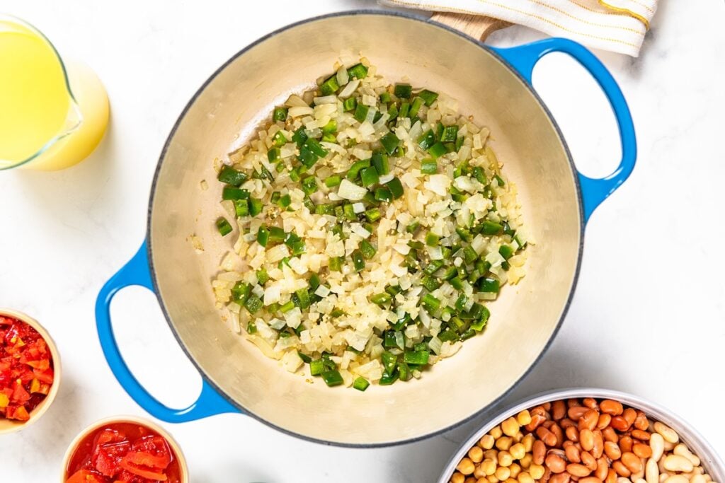 So thick and hearty, this 5 Bean Chili recipe is made in just one-pot with canned beans and a few simple pantry staples. Ready in 30 minutes! A blue enameled Dutch oven containing sautéed diced onions and diced jalapeño, surrounded by bowls of chopped tomatoes, beans, and a pitcher of broth.