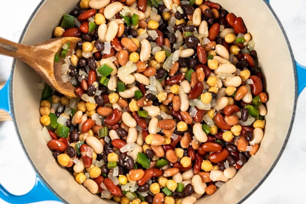 So thick and hearty, this 5 Bean Chili recipe is made in just one-pot with canned beans and a few simple pantry staples. Ready in 30 minutes! A wooden spoon stirs a mixture of black beans, chickpeas, kidney beans, and white beans with diced jalapeño and onions in a large pot.