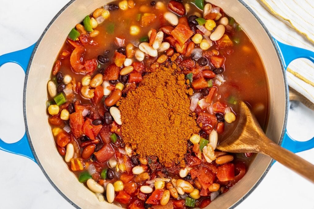 So thick and hearty, this 5 Bean Chili recipe is made in just one-pot with canned beans and a few simple pantry staples. Ready in 30 minutes! A pot of bean and tomato chili ingredients with a wooden spoon and a mound of orange spices on top.