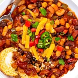 A bowl of bean chili topped with shredded cheddar cheese, diced red peppers, jalapeño slices, and cilantro, served with a piece of cornbread and a spoon.
