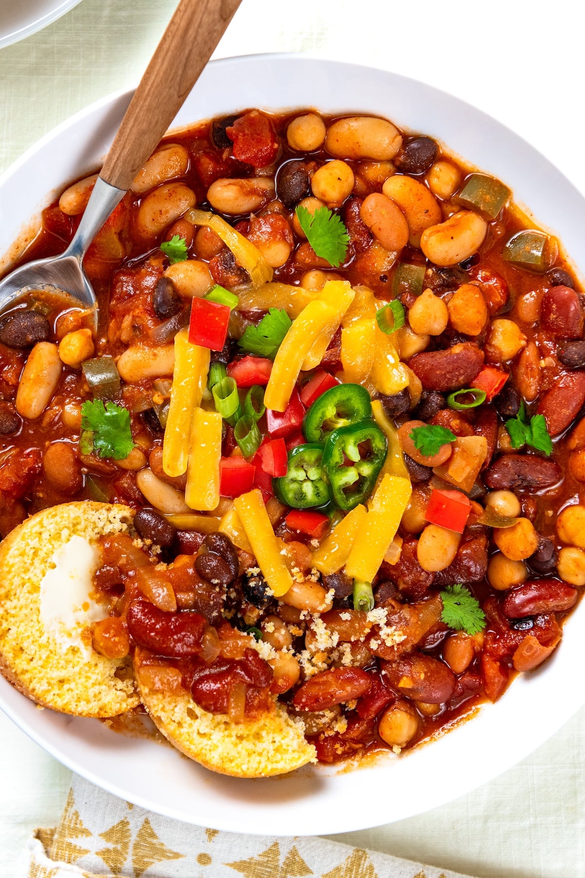 So thick and hearty, this 5 Bean Chili recipe is made in just one-pot with canned beans and a few simple pantry staples. Ready in 30 minutes! A bowl of bean chili topped with shredded cheddar cheese, diced red peppers, jalapeño slices, and cilantro, served with a piece of cornbread and a spoon.