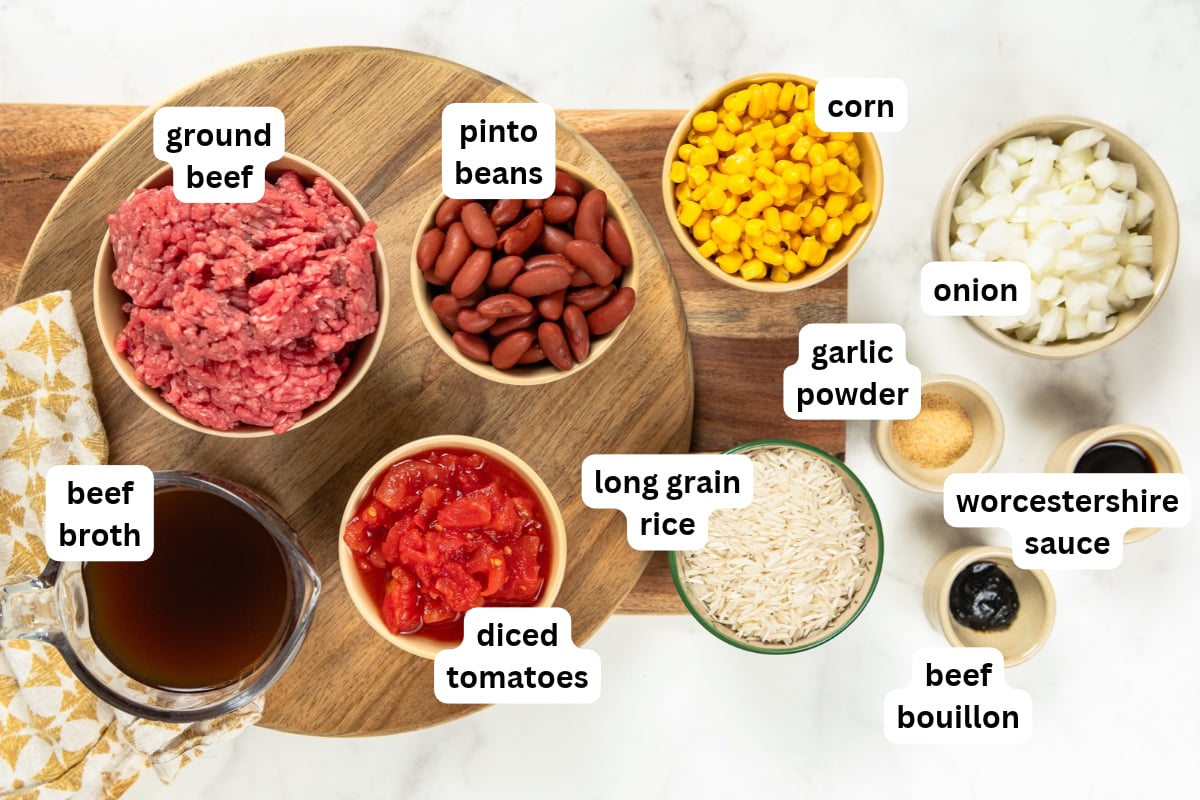 This easy Skillet Beef and Rice recipe is a hearty one-pan recipe that needs no sides. It's a budget dinner the whole family will love! Top-down view of ingredients for a skillet beef and rice recipe, labeled: ground beef, pinto beans, corn, diced onion, beef broth, diced tomatoes, long grain rice, garlic powder, Worcestershire sauce, and beef bouillon.