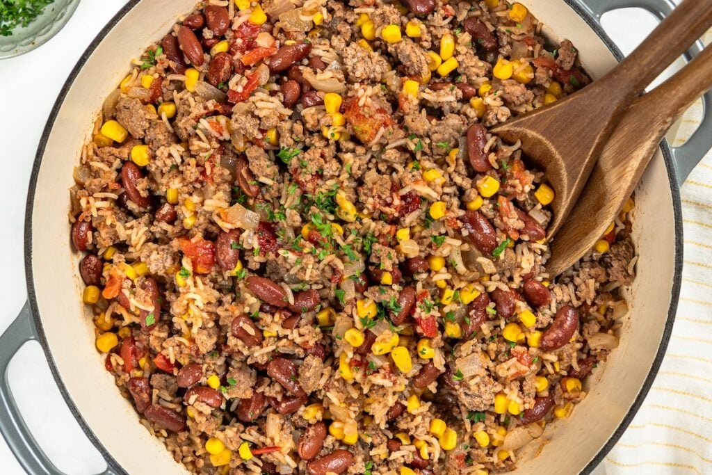 This easy Skillet Beef and Rice recipe is a hearty one-pan recipe that needs no sides. It's a budget dinner the whole family will love! A large skillet of ground beef and rice with beans and corn garnished with parsley, with two wooden spoons resting inside.