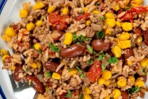 A serving of skillet beef and rice with beans and corn topped with chopped parsley, on a white plate with a fork on the side.