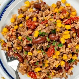 A serving of skillet beef and rice with beans and corn topped with chopped parsley, on a white plate with a fork on the side.