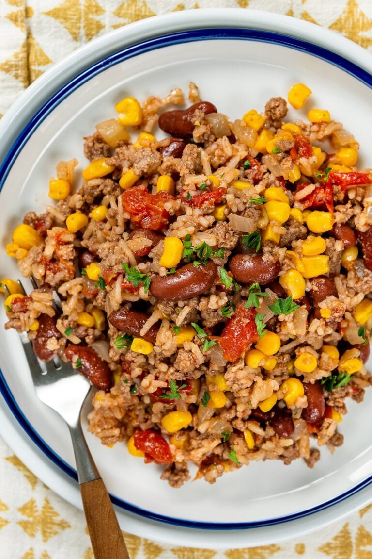 A serving of skillet beef and rice with beans and corn topped with chopped parsley, on a white plate with a fork on the side.