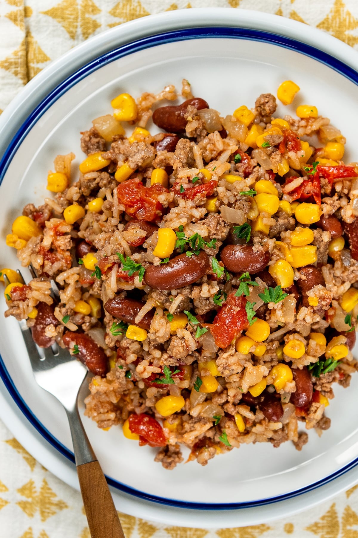 This easy Skillet Beef and Rice recipe is a hearty one-pan recipe that needs no sides. It's a budget dinner the whole family will love! A serving of skillet beef and rice with beans and corn topped with chopped parsley, on a white plate with a fork on the side.